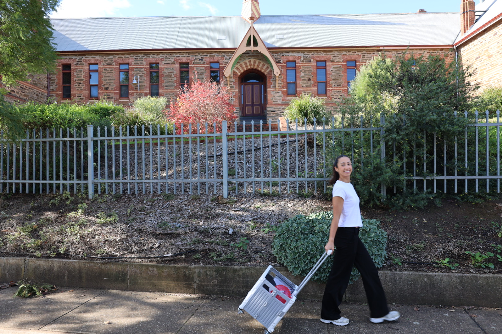 Tamsyn walking past a heritage stone schoolhouse, pulling her therapy equipment trolley